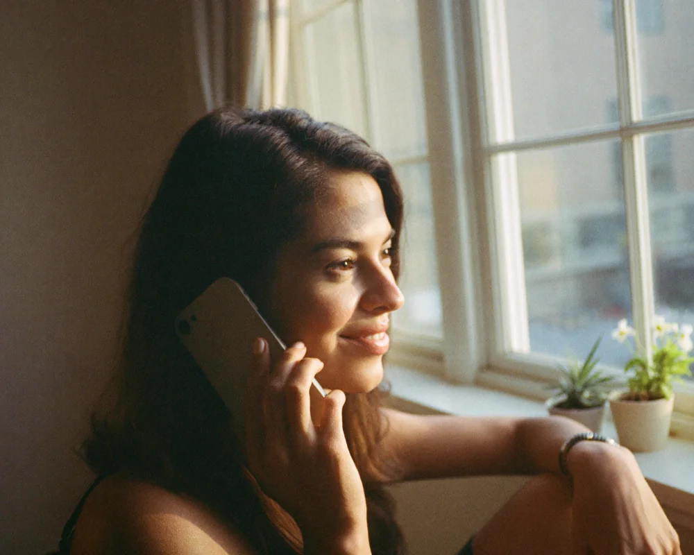 A woman smiling gently as she talks on the phone by a sunlit window